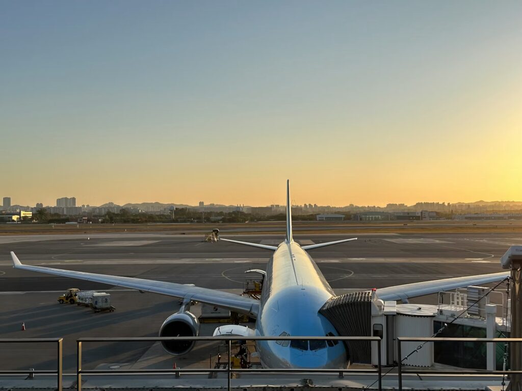 A passenger airplane parked at an airport gate, viewed from behind at sunset with a hazy city skyline in the distance.
