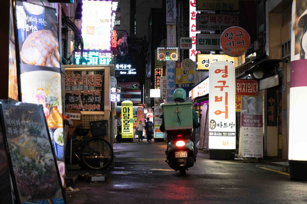 Delivery scooter riding through a neon-lit alley of restaurants at night in Seoul, South Korea