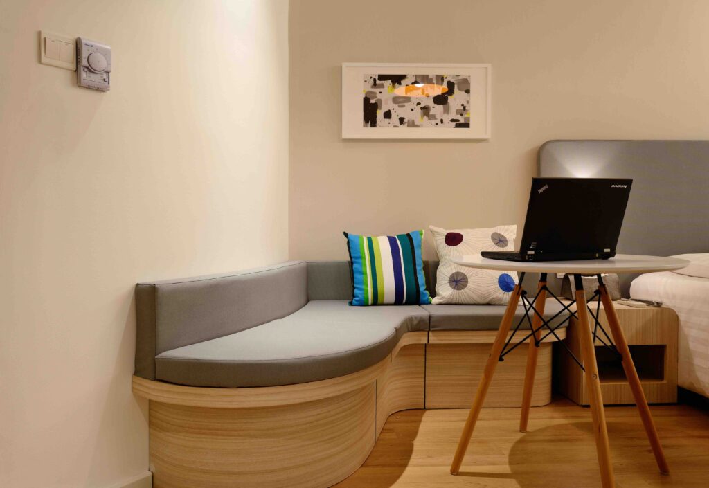 Laptop on a small table beside a hotel room bed and cushioned corner seat, set up for remote work