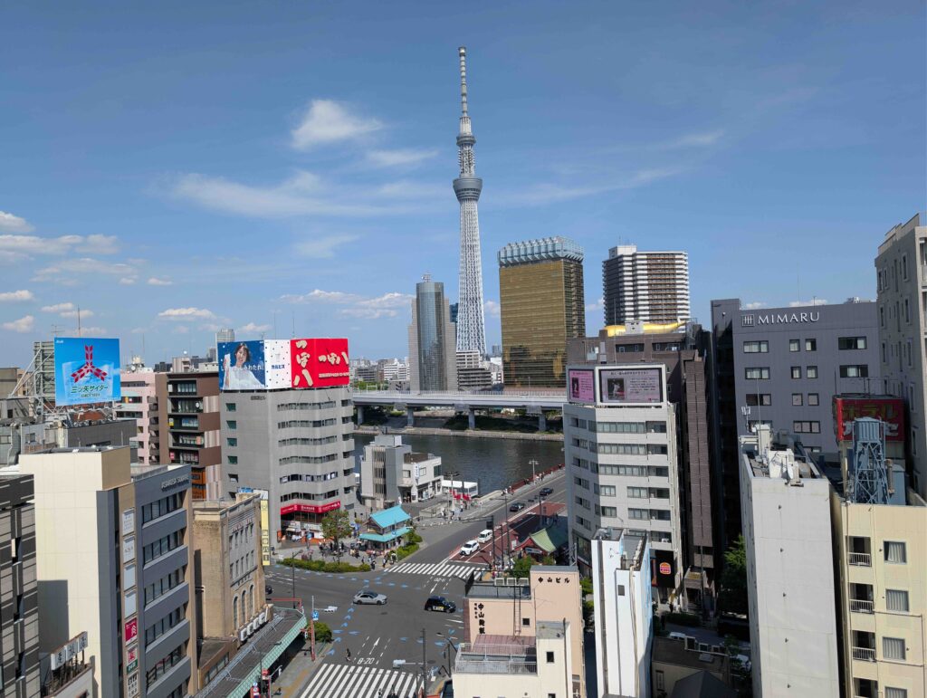 A daytime view of the Tokyo Skytree towering over a vibrant cityscape with a river, streets, and modern buildings.