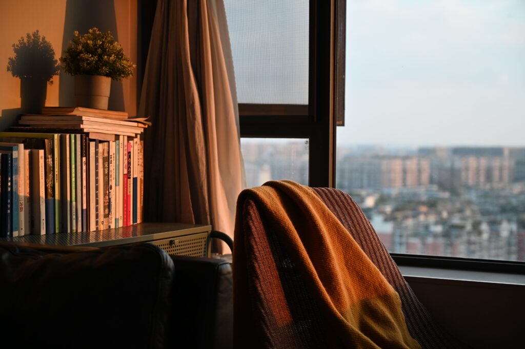 Cozy chair with a blanket by a window overlooking a city at sunset, beside a full bookshelf