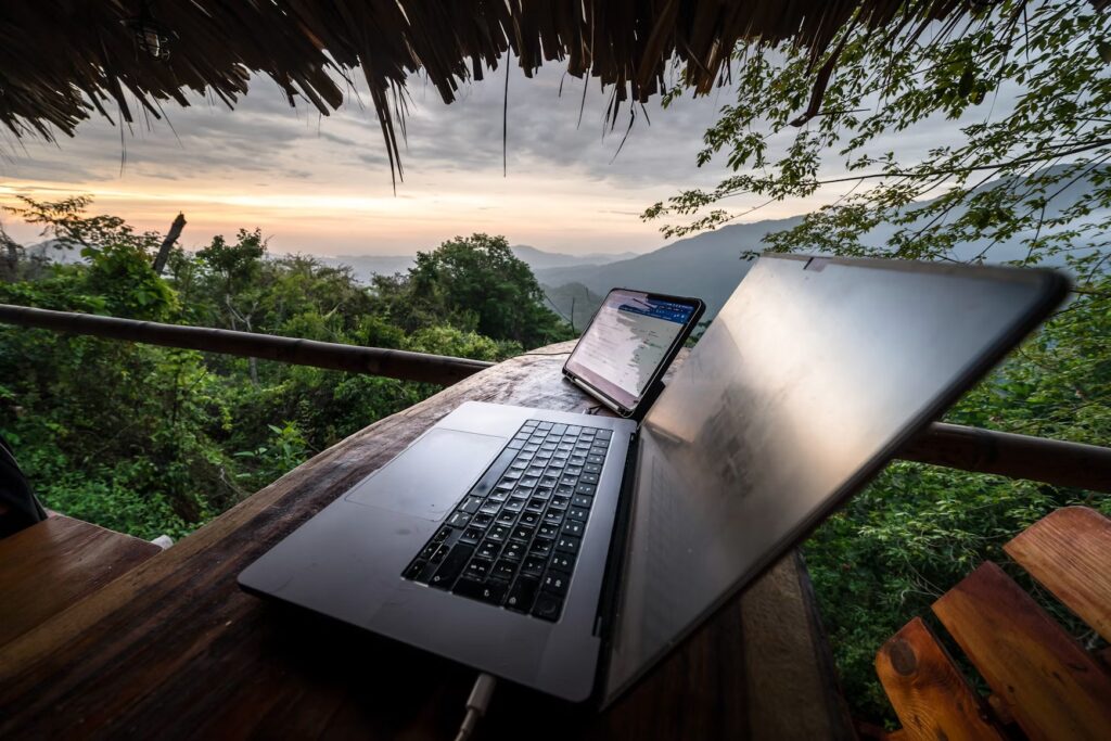 Open laptop and tablet on a wooden table under a thatched roof overlooking jungle hills at sunset