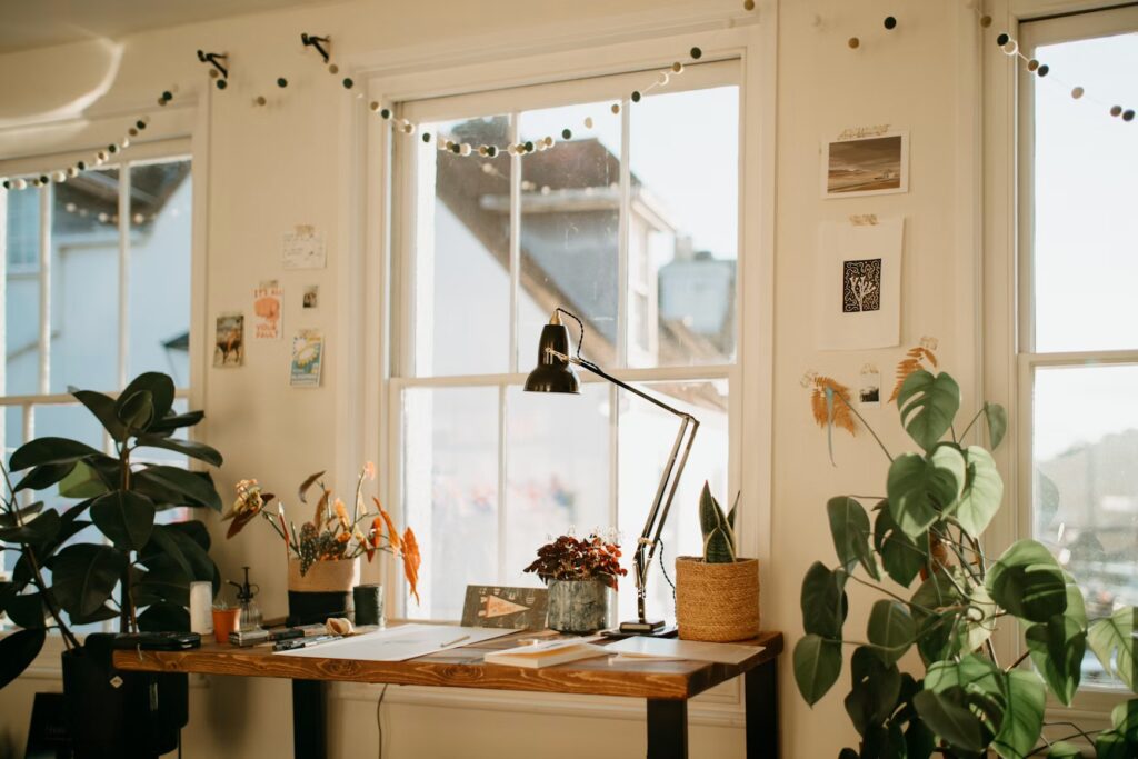 Sunlit home workspace by a window with a wooden desk, lamp and houseplants in a cozy room