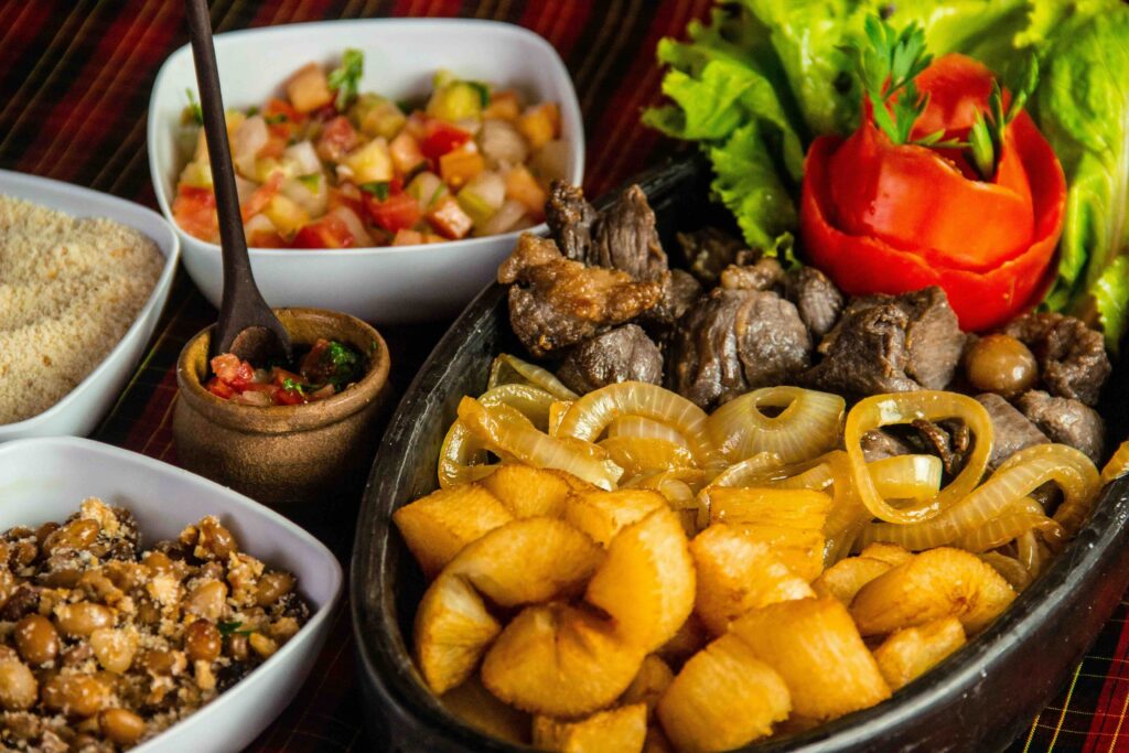 Discover Must-Try Dishes and Top Culinary Experiences on a Brazil Food Adventure A platter of Brazilian-style steak, fried cassava, and sautéed onions is served alongside sides of beans, farofa, and salsa.