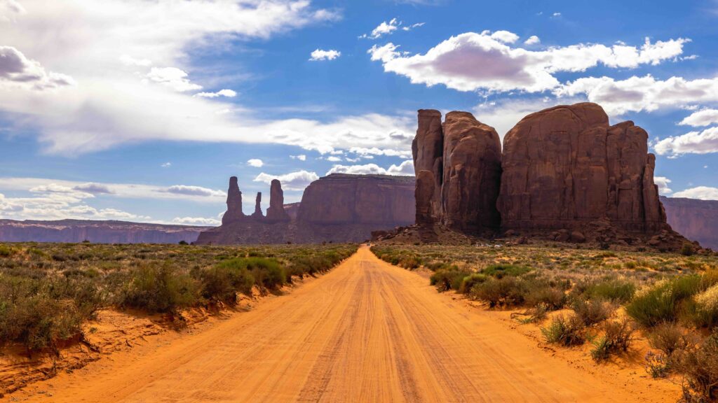 Exploring the American Southwest: Must-See Destinations, Scenic Road Trips, and Adventure Ideas A dirt road stretches toward massive red sandstone rock formations and mesas under a bright blue sky with fluffy white clouds.