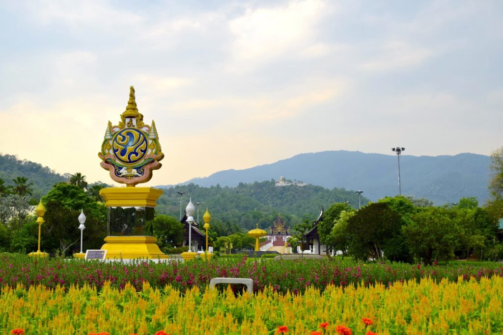 Flying from Bangkok to Chiang Mai: The Fast and Easy Way to Explore Northern Thailand A golden Thai emblem monument surrounded by vibrant flowers and lush green mountains under a cloudy sky.