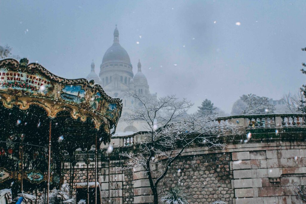 Christmas in Paris (2026): 25 Magical Things to Do (Including Free Activities) Snow falling over a vintage carousel with the Sacré-Cœur Basilica in the background.