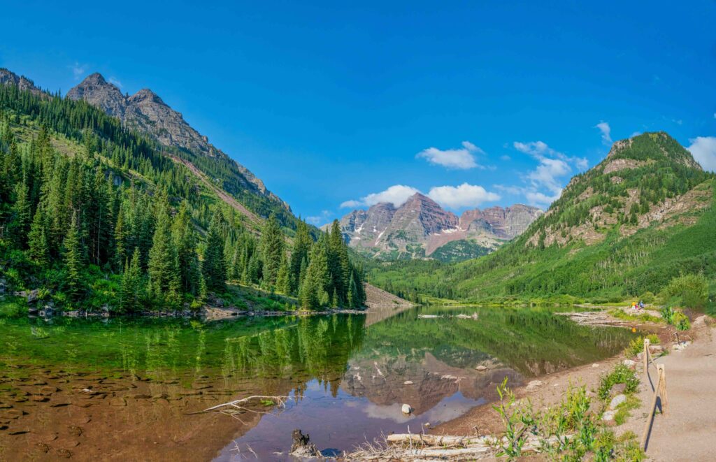 Lush green pine trees and the Maroon Bells reflecting in the clear lake water. 