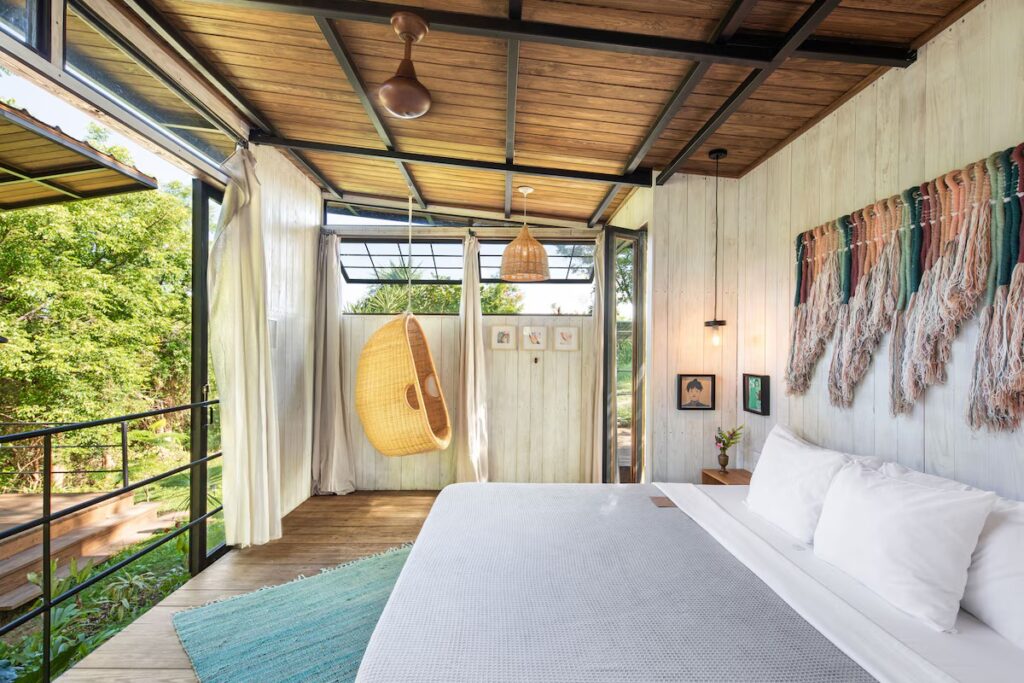 Minimalist, nature-inspired bedroom with a hanging chair and open windows at Finca Victoria. 