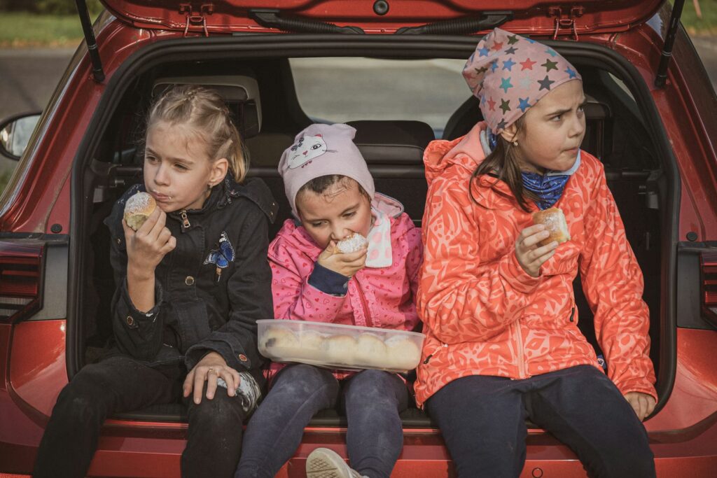 Three kids eating snacks from a plastic container in the car trunk, an essential of the [family travel hacks] to prevent hangry meltdowns during a road trip