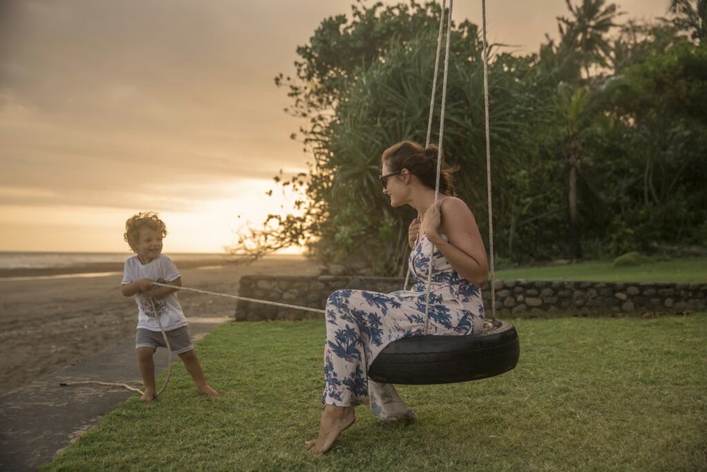 A mother and her toddler laughing while playing on a tire swing by the beach at sunset, capturing the joy of stress-free family vacations