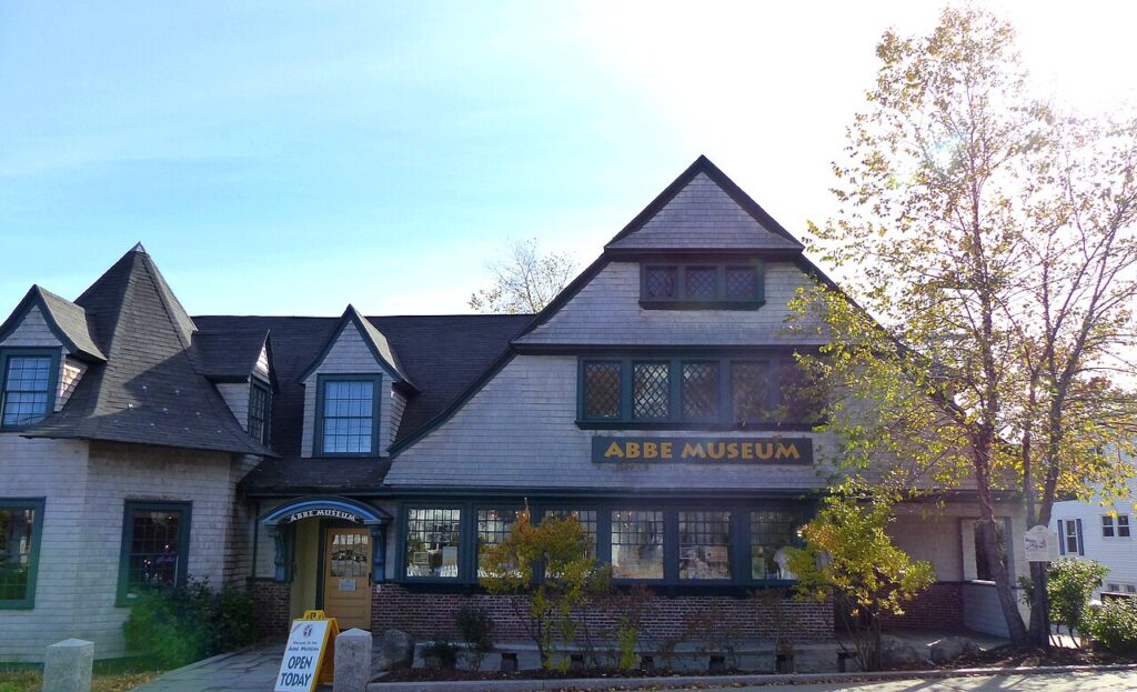 The historic, shingled exterior of the Abbe Museum building with a yellow sign on the door.