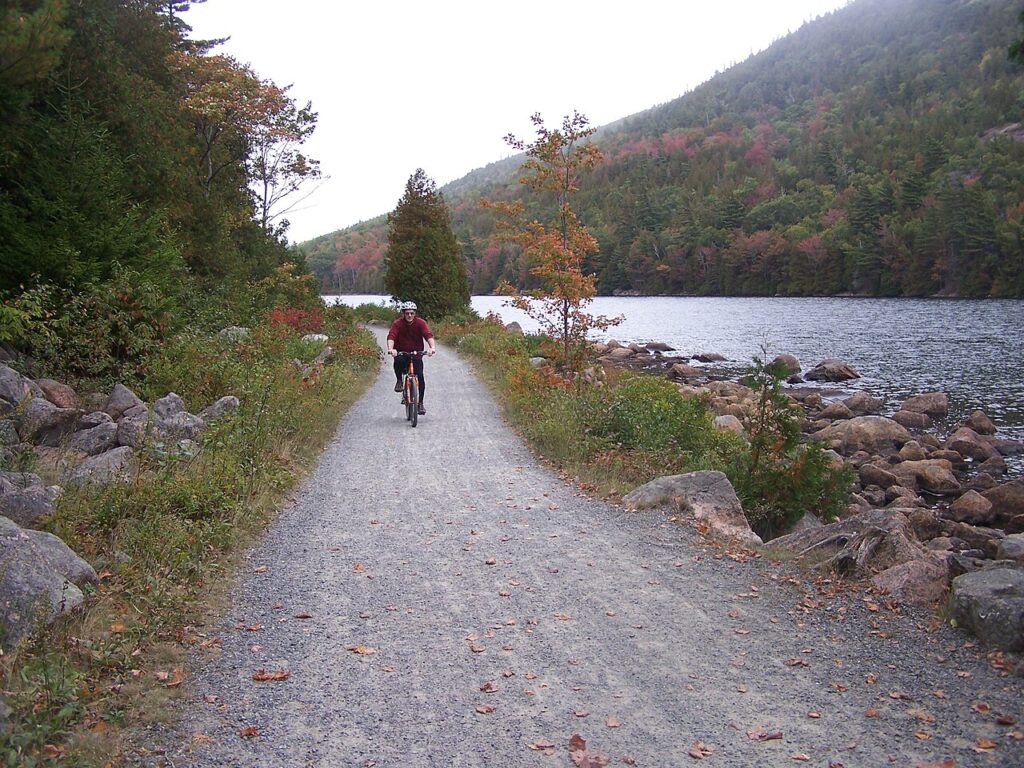 A person riding a bicycle along a wide, crushed-gravel path next to a rocky lake shoreline.