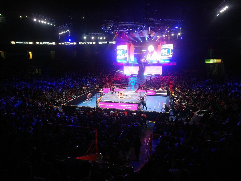 A large, energetic crowd watching an intense Lucha Libre wrestling match inside a brightly lit arena. 