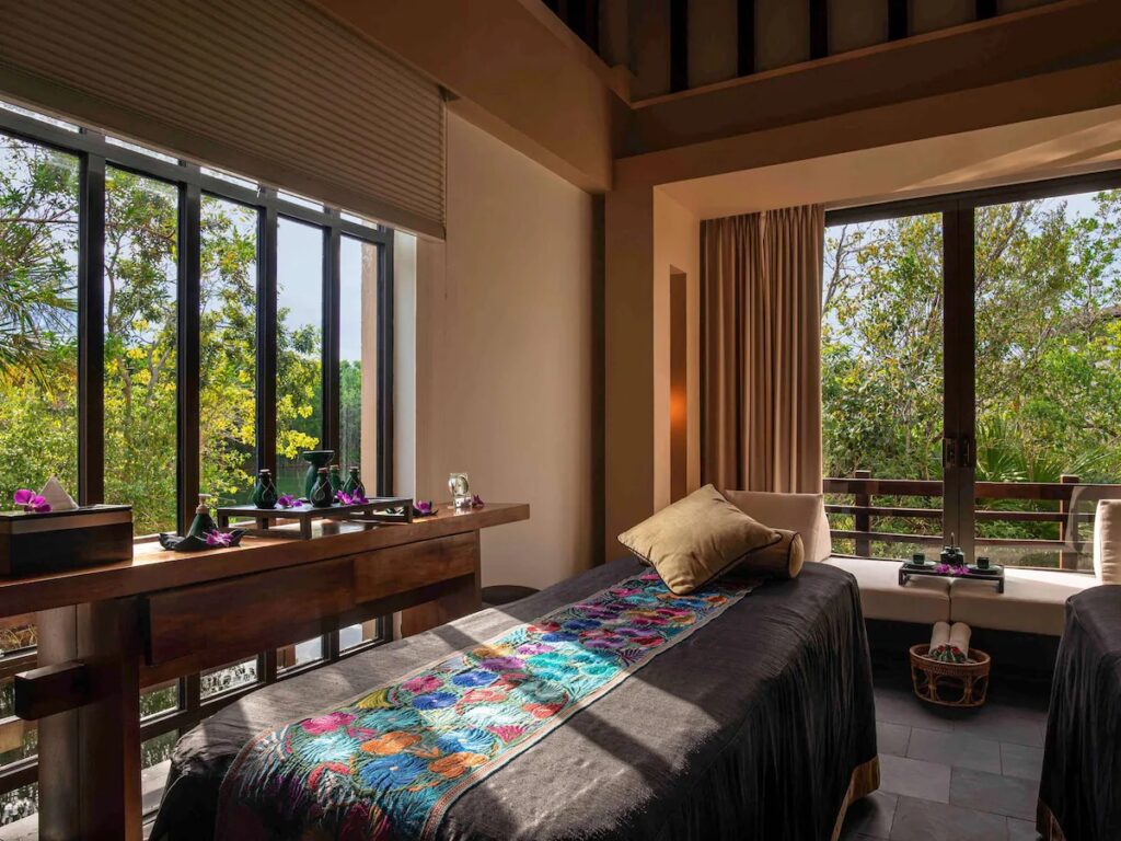 Two massage tables set in a tranquil spa treatment room with large windows facing the jungle at Banyan Tree Mayakoba.