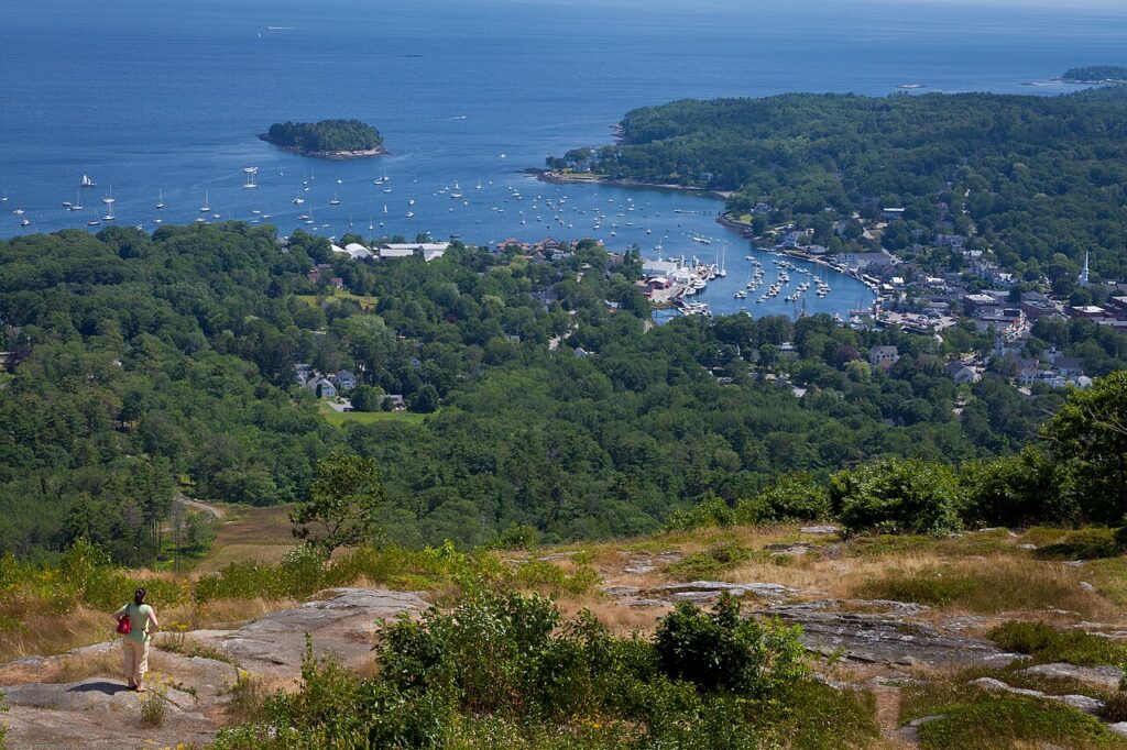 A person walking on the rocky granite summit of Mount Battie looking down at the boat-filled Camden Harbor.
