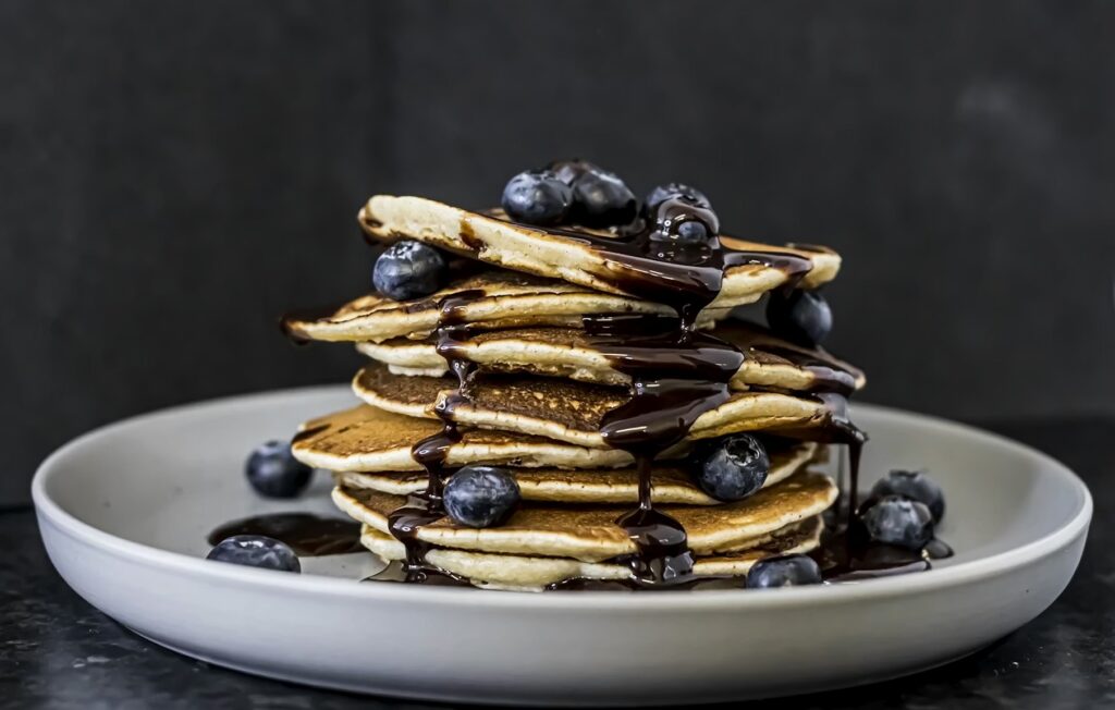 A tall, delicious stack of wild blueberry pancakes dripping with dark syrup on a white plate.