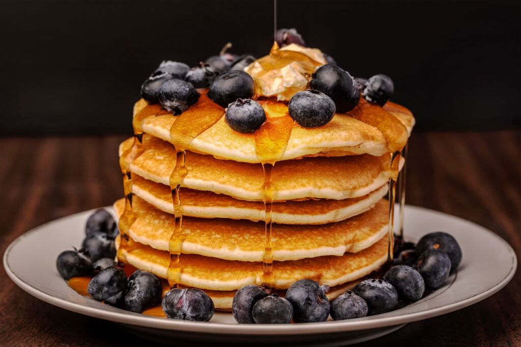 A tall stack of fluffy pancakes topped with butter, syrup, and fresh wild blueberries.