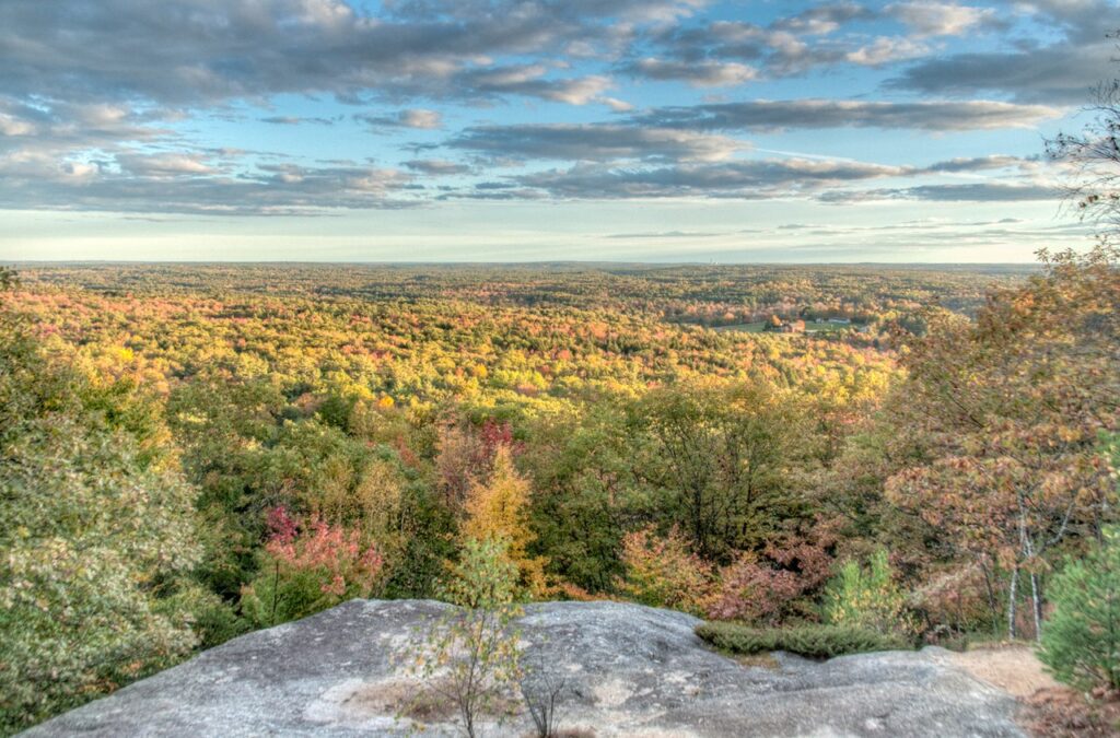 A sweeping panoramic view of a vast, colorful autumn forest from a high granite rocky ledge.