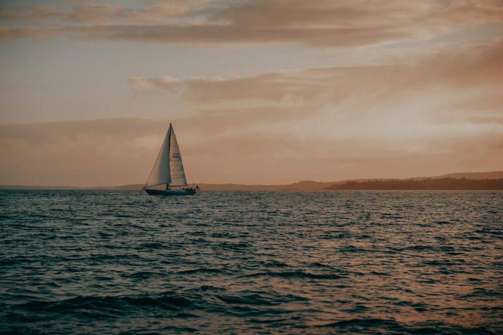 A small sailboat cruising on the calm ocean waters during a muted, golden sunset.
