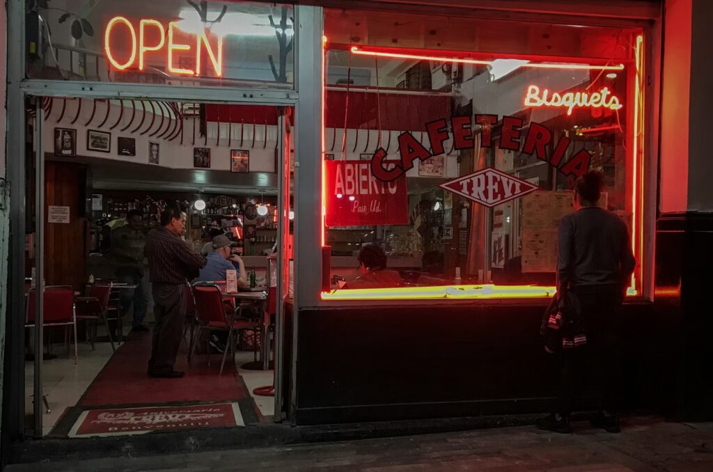 The glowing neon signs of a traditional late-night diner or cantina viewed from the street. 