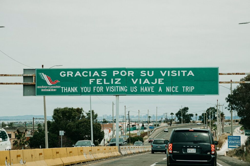A green road sign at the US-Mexico border crossing reading "Gracias por su visita" as cars depart the country.