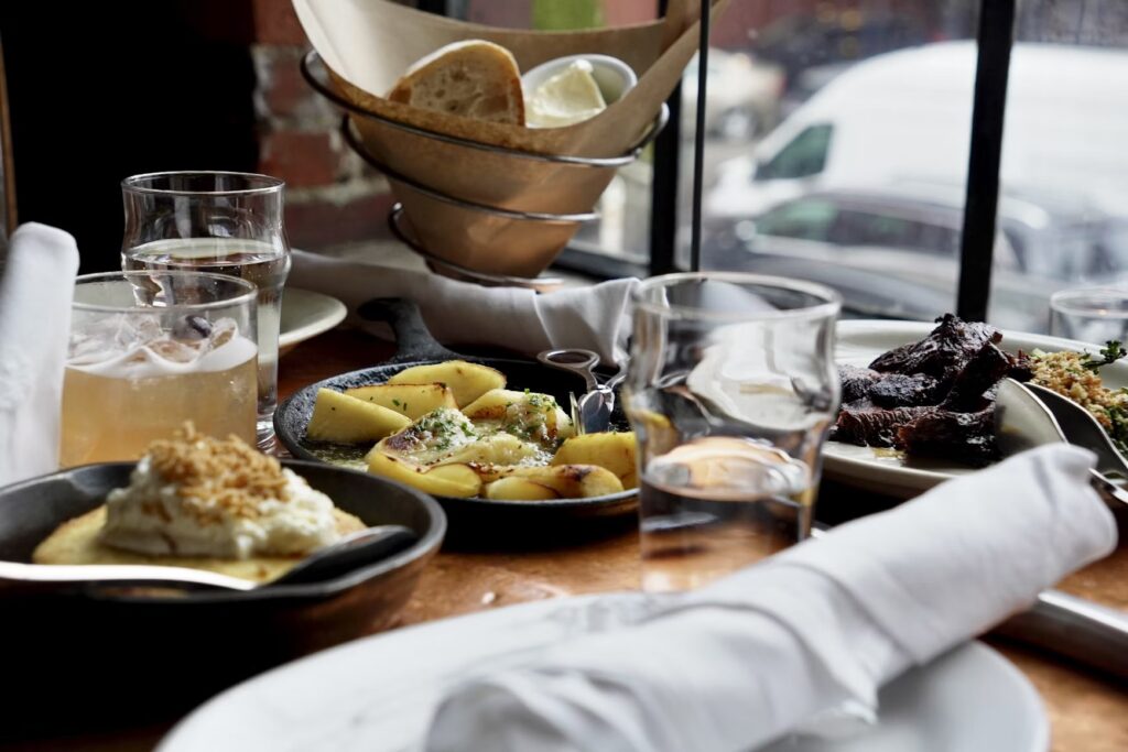 A table by a window featuring a spread of small plates, including roasted potatoes, sliced meat, bread, and drinks. 