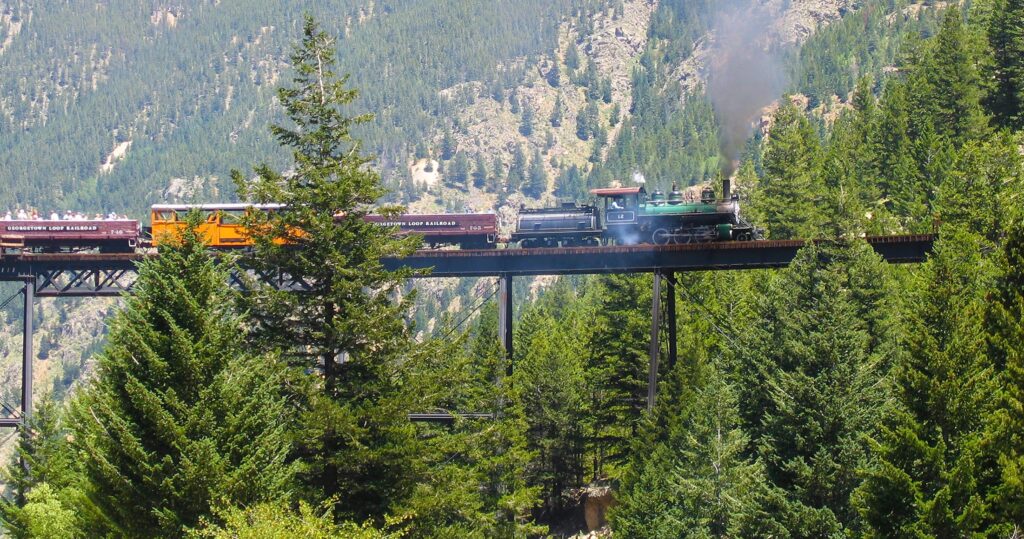 A historic steam train crossing a high trestle bridge over a green pine forest. 