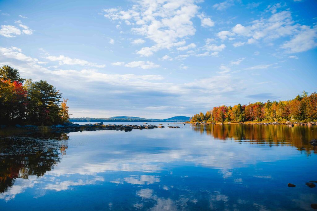 Maine in the Fall (2026): The Ultimate Road Trip & Itinerary Guide A serene lake reflecting vibrant autumn foliage and a clear blue sky in Maine, illustrating Maine in the fall.