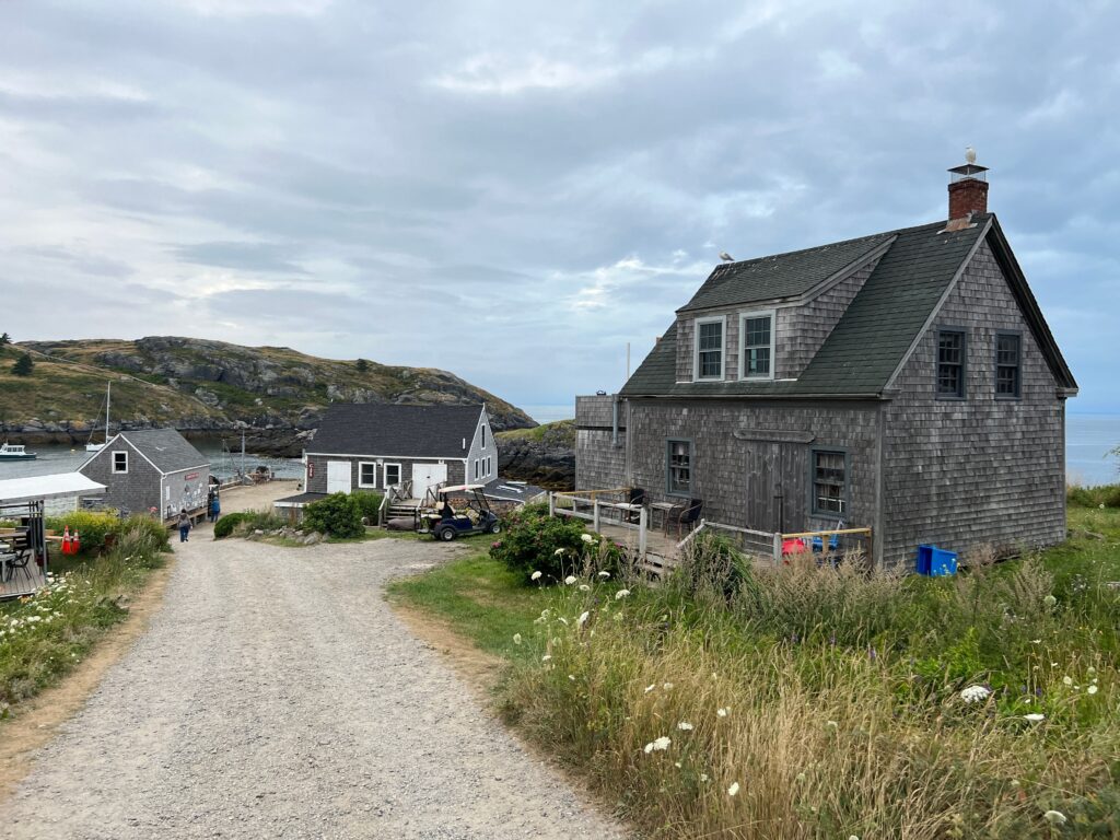A dirt path leading past weathered shingled houses toward a quiet island harbor.