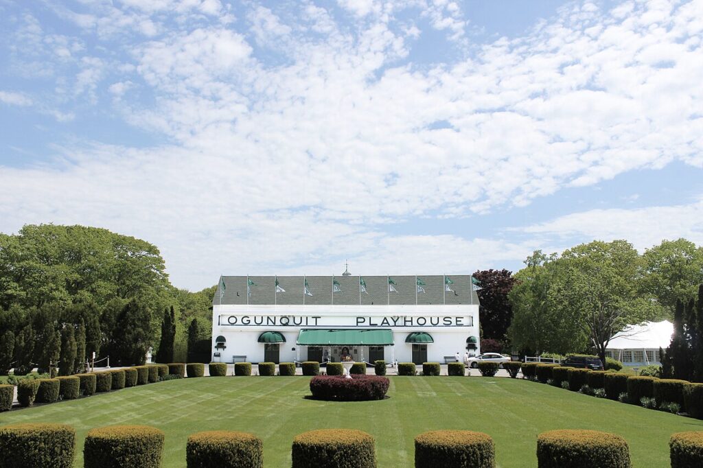 The historic white exterior of the Ogunquit Playhouse building sitting behind a manicured green lawn in Maine.