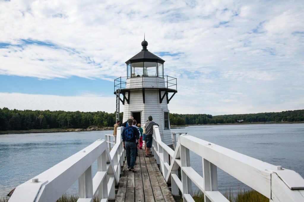 Visitors walking along a wooden pier toward a classic white lighthouse positioned over the water.