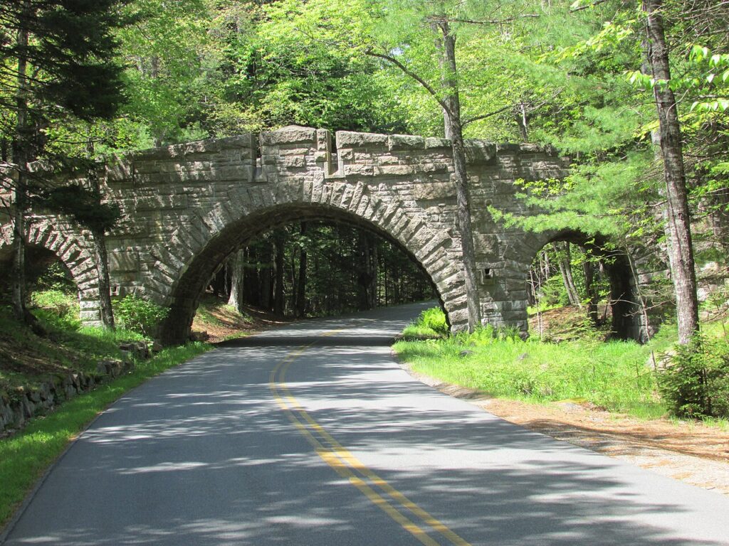 A historic, arched stone bridge spanning a paved road surrounded by green summer trees.