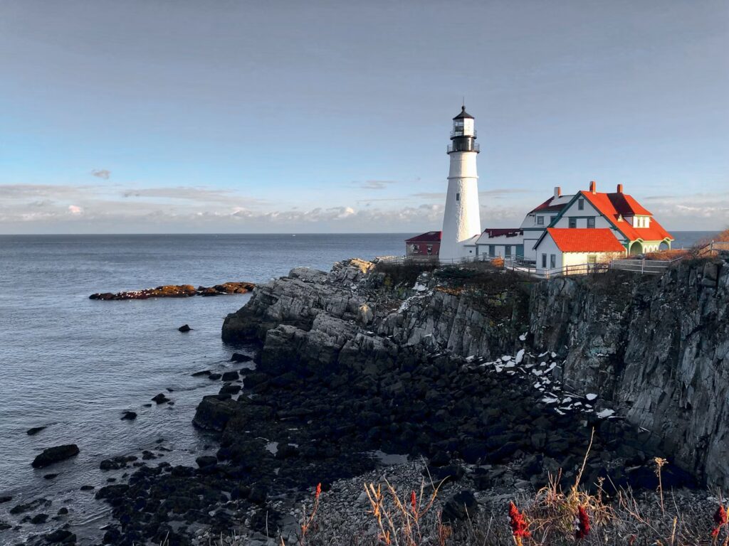A beautiful white lighthouse and keeper's quarters standing on a rugged, snow-dusted cliff by the ocean. 