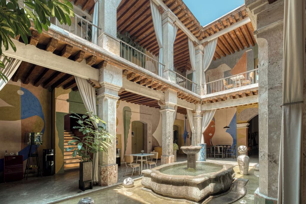 A stunning, sunlit interior courtyard featuring painted wall murals and a stone fountain at Pug Seal Oaxaca.
