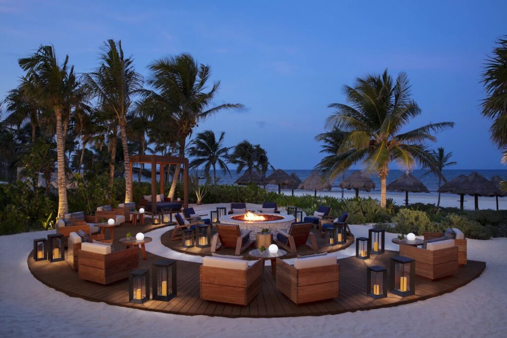 Cozy outdoor fire pit lounge set on the sand at dusk at Secrets Maroma Beach. 