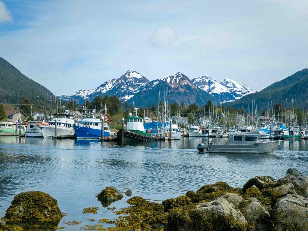 Fishing boats docked in a calm harbor with snow-capped mountain peaks in Sitka, Alaska.