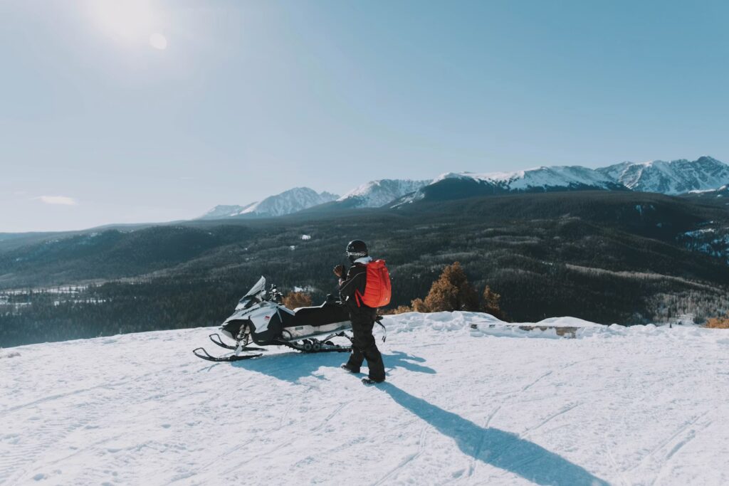A person standing next to a parked snowmobile on a snowy mountain ridge. 