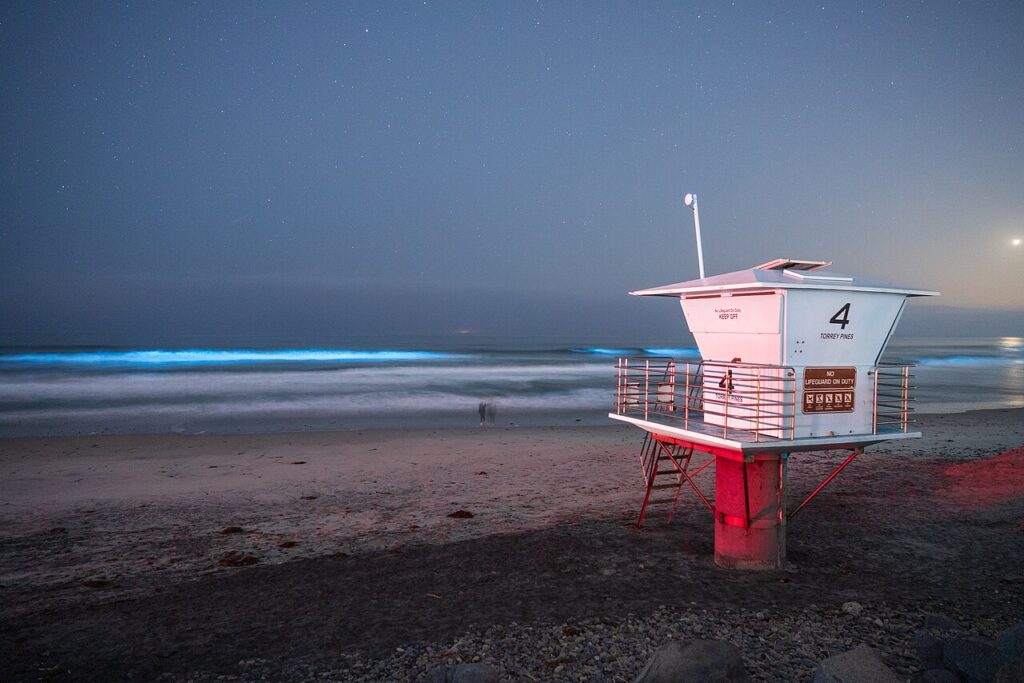 Vivid blue waves crashing near a white lifeguard tower, highlighting the Bioluminescence California phenomenon.