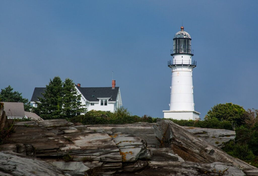 A tall white lighthouse and historic keeper's house overlooking rocky coastal ledges under a blue sky. 