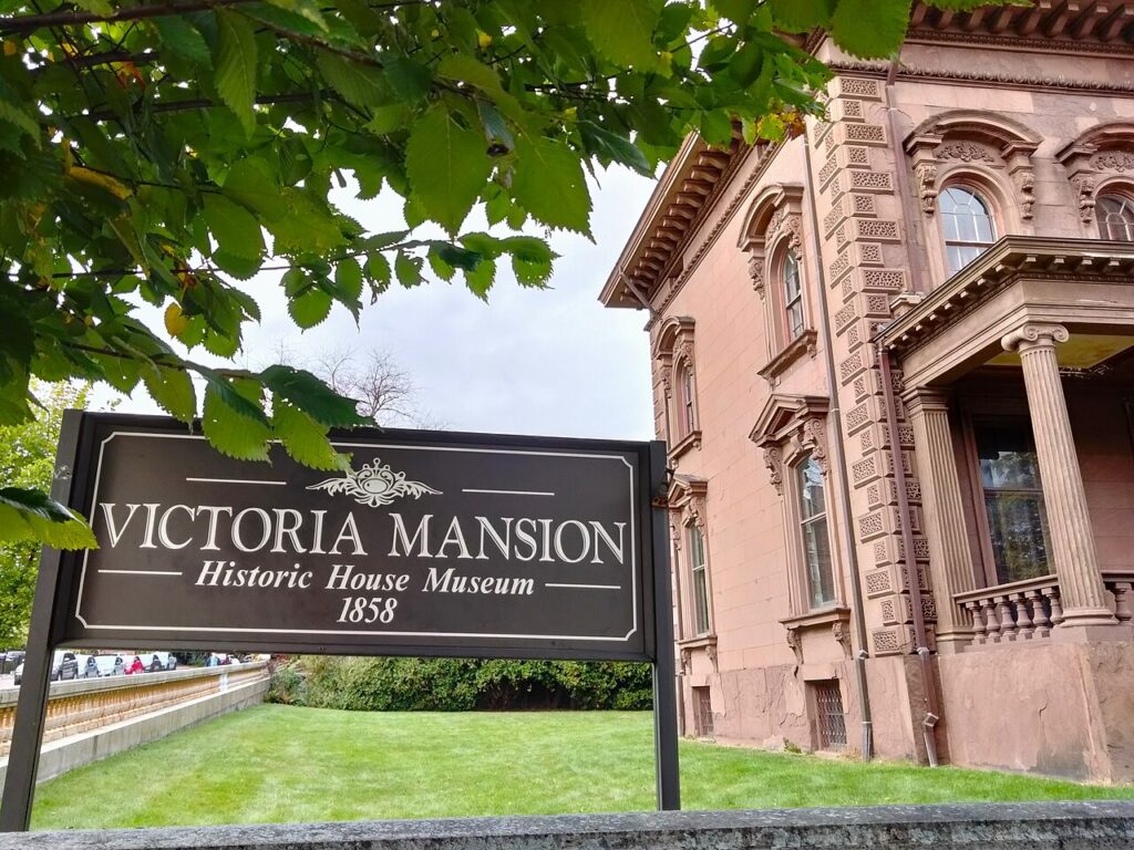 The Victoria Mansion Historic House Museum sign standing on the lawn in front of the pre-Civil War building. 