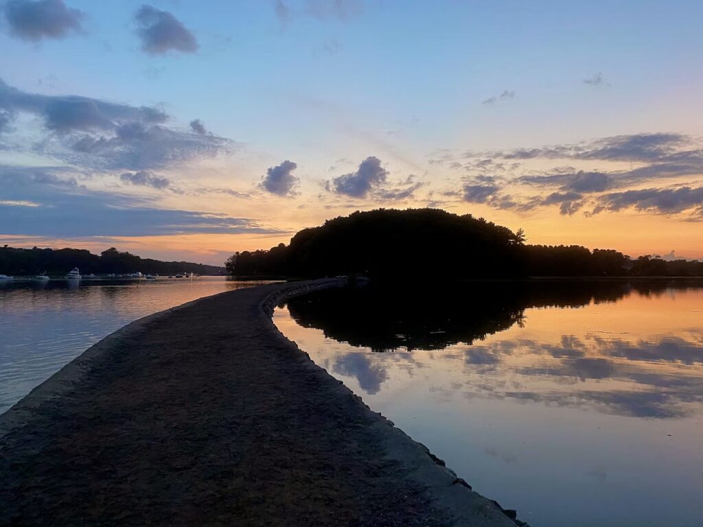 A sunset view of the narrow path leading to the tiny Wiggly Bridge across a calm tidal channel in York, Maine.
