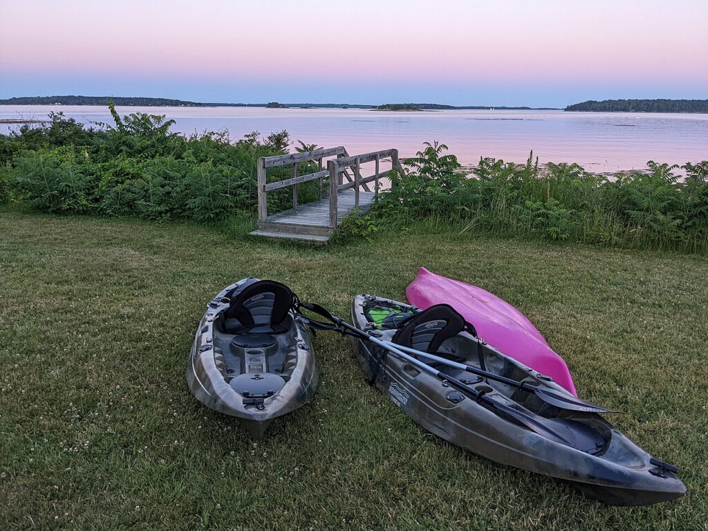 Two kayaks resting on the grassy shore of a calm bay during a soft, colorful sunset.