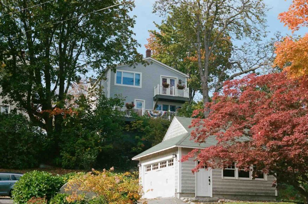 A quaint two-story house with a white garage perched above a red autumn tree in Wiscasset, Maine.