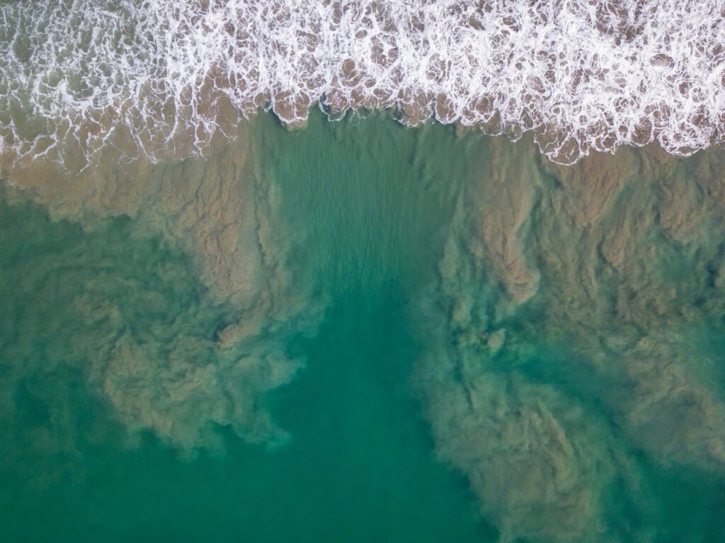 Aerial view of a reddish-brown red tide mixing in the ocean waves along the San Diego coast during the day.