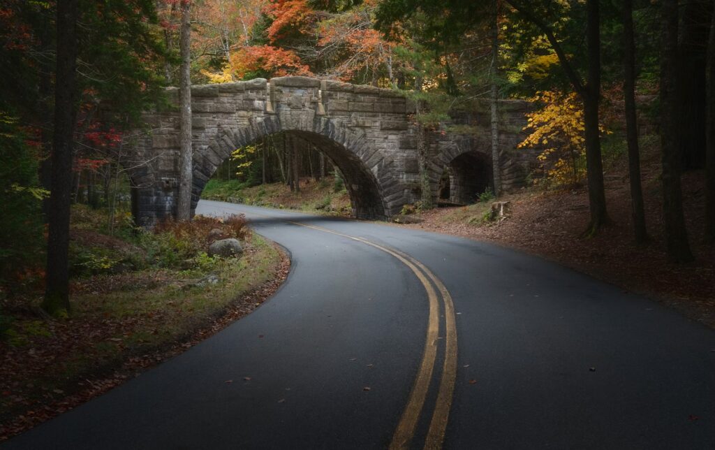 A classic arched stone bridge crossing a winding paved road surrounded by colorful autumn foliage.