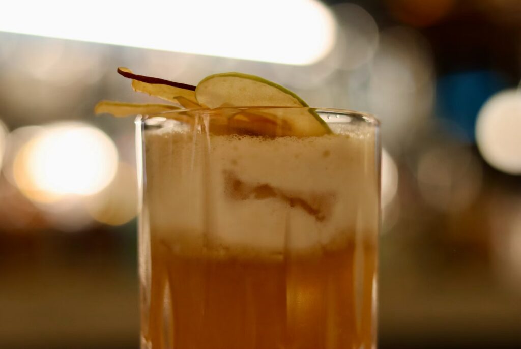 A close-up of a beautifully crafted speakeasy cocktail topped with foam and a delicate apple slice garnish. 