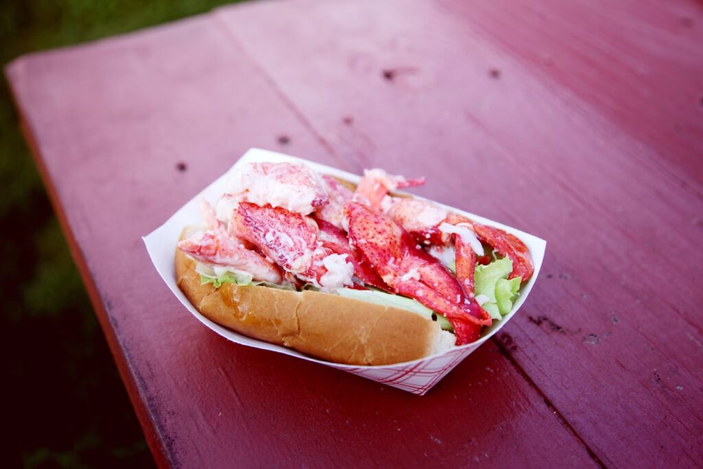 A fresh, delicious Maine lobster roll served in a paper tray on a rustic red picnic table in Perkins Cove.