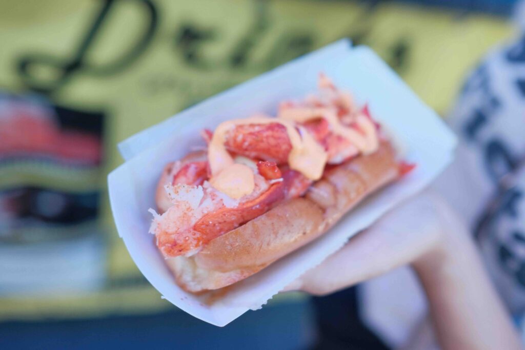 A person holding a delicious lobster roll topped with a creamy sauce in a paper tray from Bite Into Maine. 