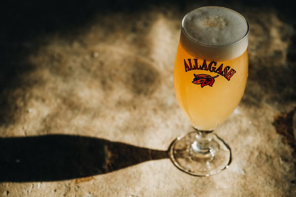 A glass of Allagash craft beer glowing on a textured surface under a dramatic spotlight.